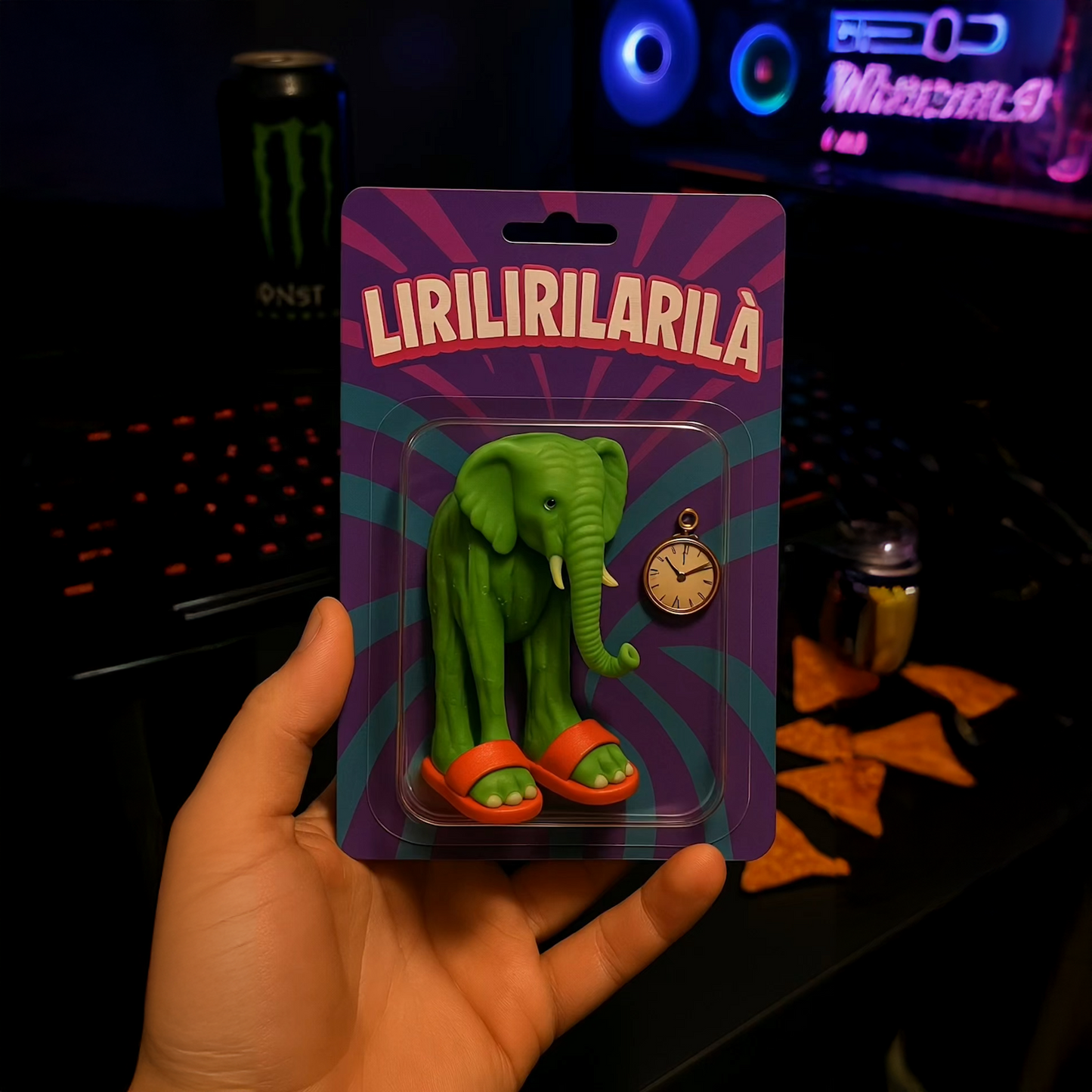 Green elephant plush toy named Lirilirilarilà in retro blister packaging, wearing red slippers and holding a pocket watch, in front of a gaming setup with Monster and chips.